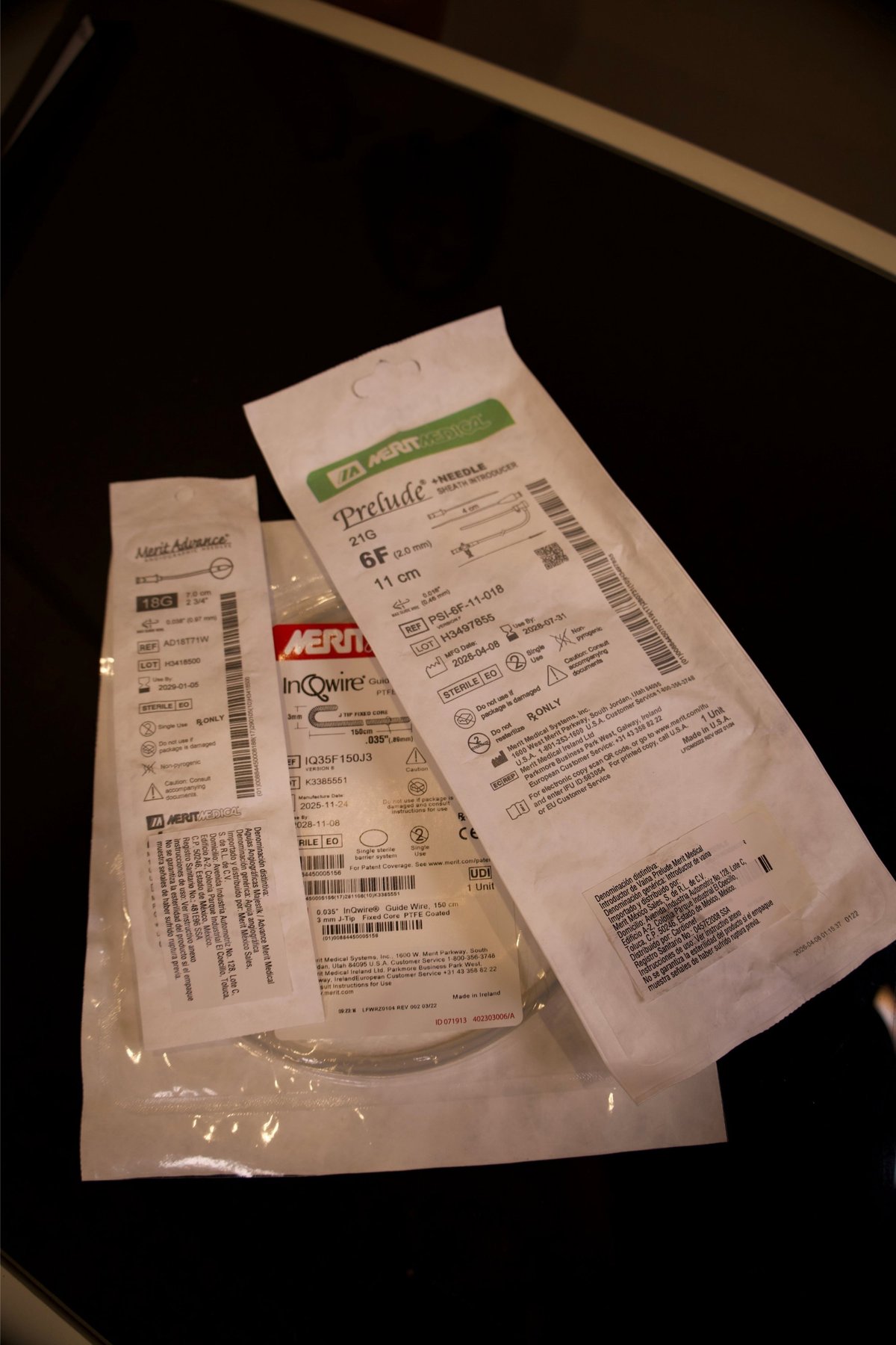 Etiquetas y empaques de dispositivos Merit Medical — Merit Advance 18G, InQwire guidewire y Prelude 21G sheath — sobre superficie oscura con información de empaque visible